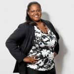 A black woman with a sleek ponytail poses with her hands on her hips staring directly at the camera. We can see her outfit from the hips up: an abstract black-and-white-spotted blouse and a black blazer