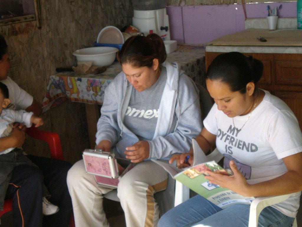 Two people lounge in plastic chairs in casual clothing. The person on the right in focused on a box in their hands; the person on the right is intently leafing through a guide about community banks