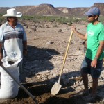 Two men stand in front of an arid plateu, one of them holding a shovel