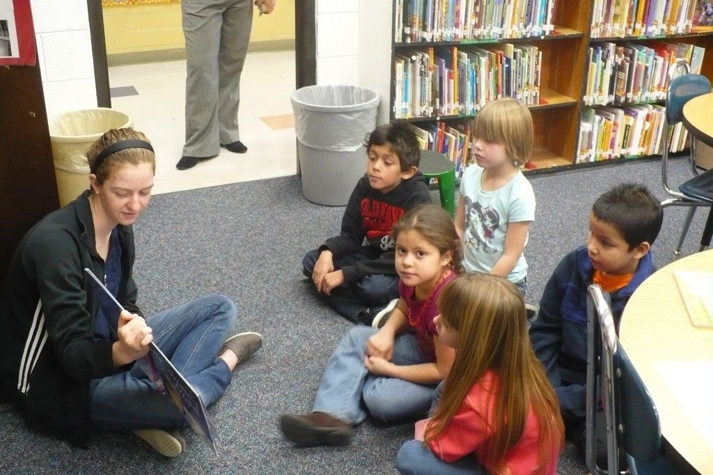 A teenager sitting on the ground reads a book to 5 children, one of whom is looking at the camera.