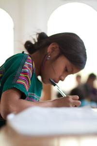 a child with dark hair and a bright emerald tunic hunches over schoolwork, writing