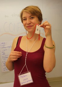 A person in a red tank top stand smiling in front of a whiteboard. They are wearing a lanyard nametag and holding up a slip of paper with the letter "e" on it