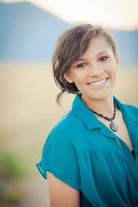 A professional portrait of Aleiya where she is shown smiling in front of a dramatically blurred background. Her wavy brown hair is swept into a ponytail and she is wearing a vibrant blue blouse