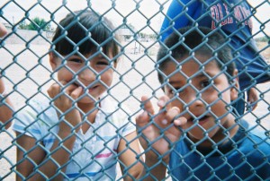 Two children grasp the links of a wire fence. the photo is taken from the other side