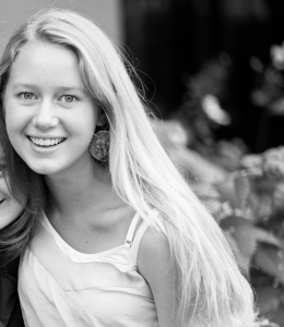 Nikola leans against someone who has been cropped out of the photo. She has straight, chest-length blonde hair, pale skin, and a white tank top. The photo is black and white with a dramatically blurred background