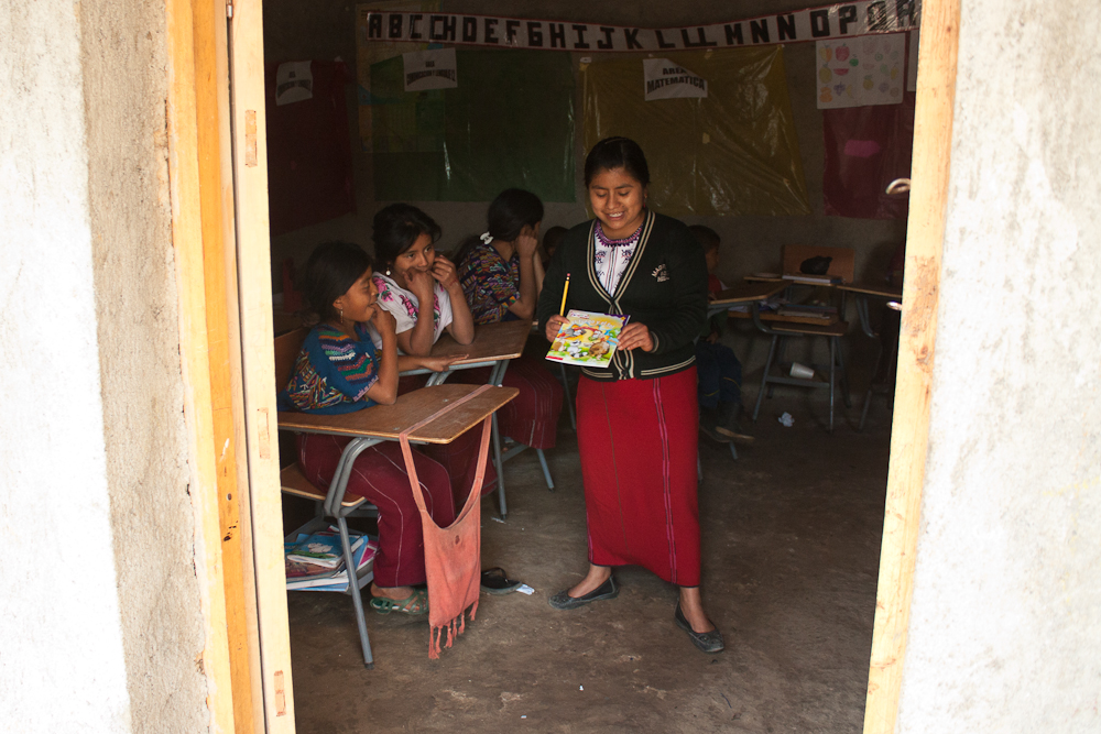 a photo taken through an entryway reveals three children at school desks and one standing up with a book in their hands