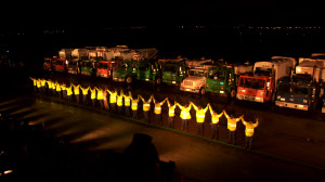 a line of dozens of people stand holding raised hands in front of a line of trucks