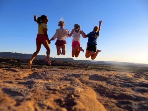 four teenagers are pictured mid-jump against a bright blue sky and desert ground