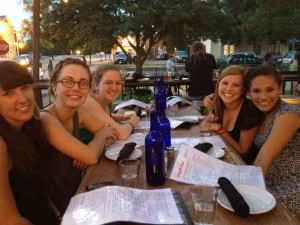 Five girls sit at an outdoor wooden dining table covered in menus, plates, and silverware rolled in navy napkins. They are looking at the camera and smiling.