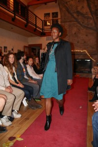 A black woman with short curly hair is pictured mid-stride down a red carpet. She is wearing a blue knee-length dress, an oversized gray blazer, and black heeled boots. In the background, we see a singular row of audience members lining the runway and a rock wall.