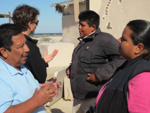 Four people stand facing each other in pairs, each of them conversing. In the background, we see stucco walls, thatched roofs, blue sky, sand, and a waterfront.
