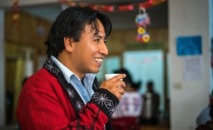 Ernesto, a man with long brown hair to the nape of his neck, a black-lined red blazer, and a styrofoam cup in his hand, smiles into the distance. The photo is in profile, taken from his right side.