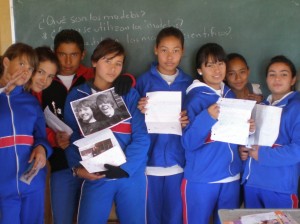 8 children in matching blue jumpsuits pose in front of a green chalkboard. Many are holding up sheets of paper or photos and smiling at the camera.