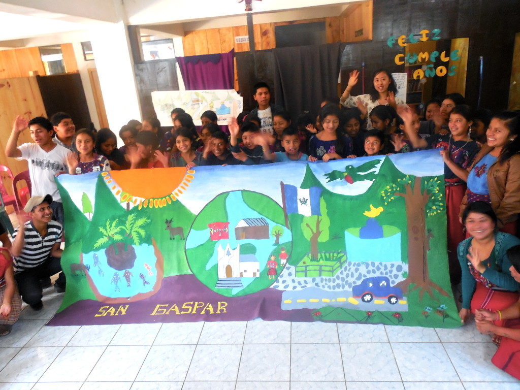 Dozens of children stand around a hand-painted banner that says "SAN GASPAR." They are in motion, waving and blurry.