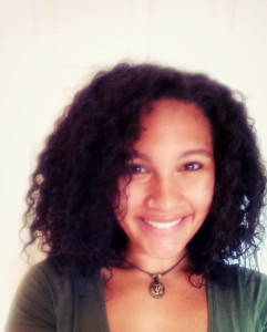 A woman smiles at the camera in a close-cropped headshot. She has curly shoulder-length hair, a nose ring, and pendent necklace.