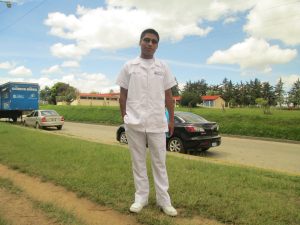 A man with dark hair and an all-white outfit poses in front of green grass and a small residential road.