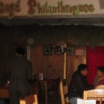 A few sets of chairs and tables, one of them occupied by a couple, are seen under a hand-painted sign that says "Philanthropiece."
