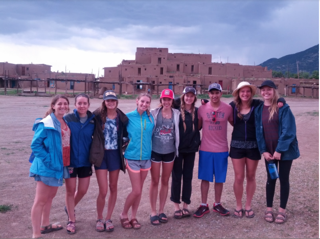 Nine people stand in a line with their arms around one another. In the background, we see a traditional pueblo structure and dark clouds.