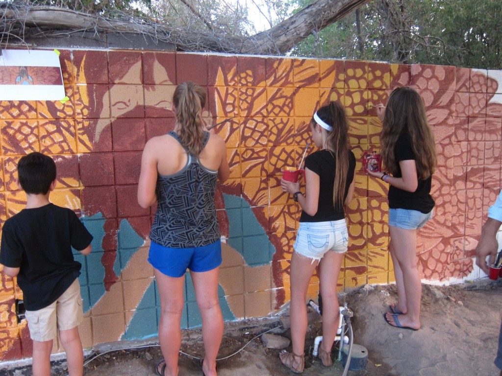Four people are pictured from behind. They are wearing sandals and shorts and painting a mural featuring lots and lots of corn.