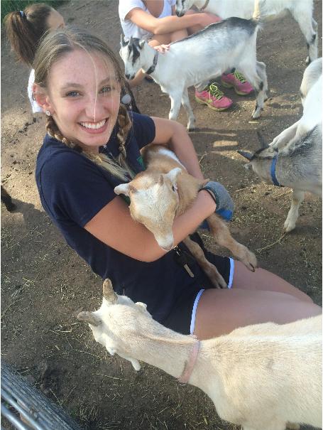 A teenager with two blonde braids kneels on dirt holding a baby goat.