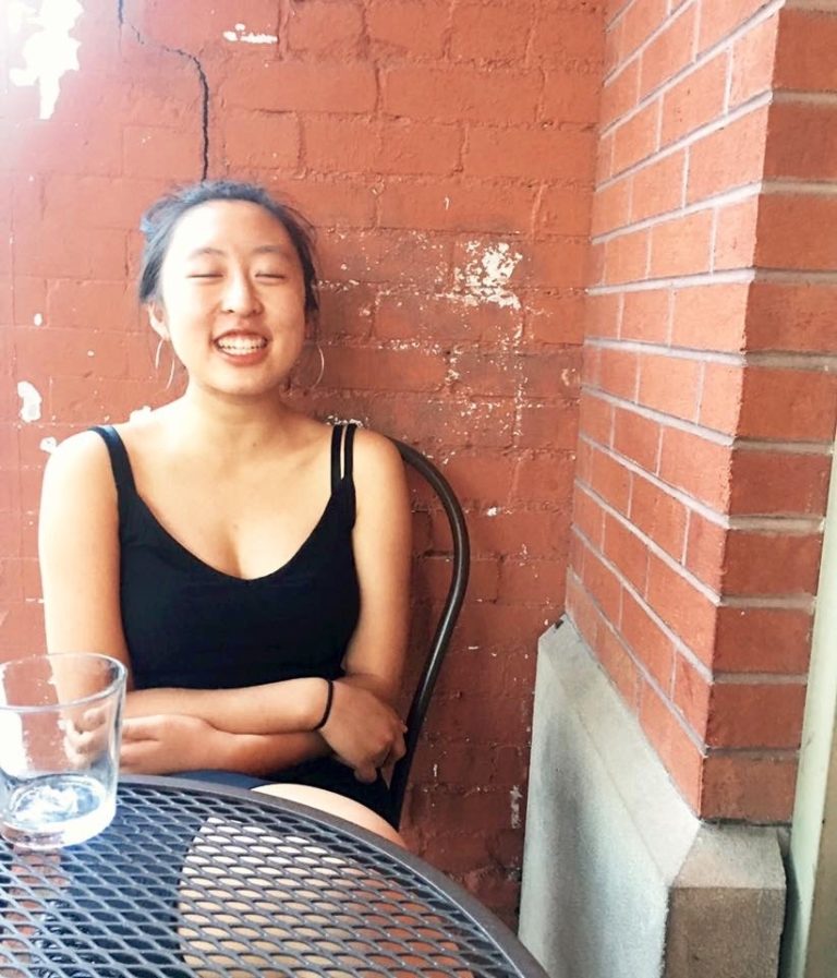 A candid portrait of Emily Sun. She has her eyes closed, laughing, and is sitting outside at a cafe table.