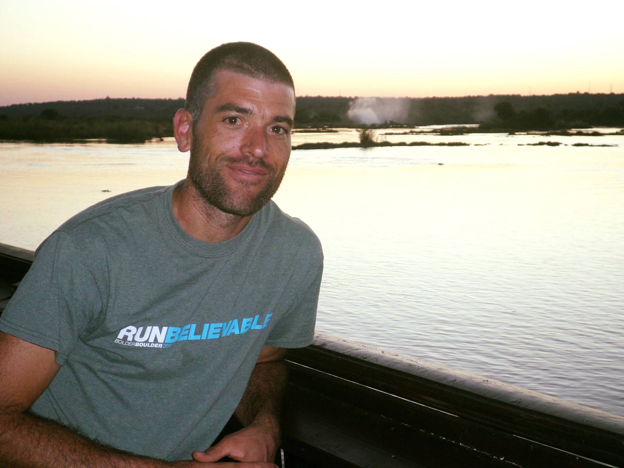 a white man with a close-shaved head and beard stands in front of a lake at dusk. He is smiling with his lips closed and wearing a "RUNbelievable" shirt