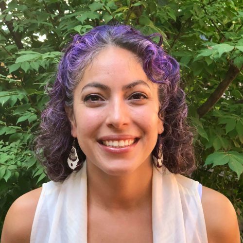 A Latina woman in a white sleeveless blouse and big dangle earrings poses in front of foliage. She has shoulder-length curly purple hair, eyeliner, and a smile that reveals her top teeth.