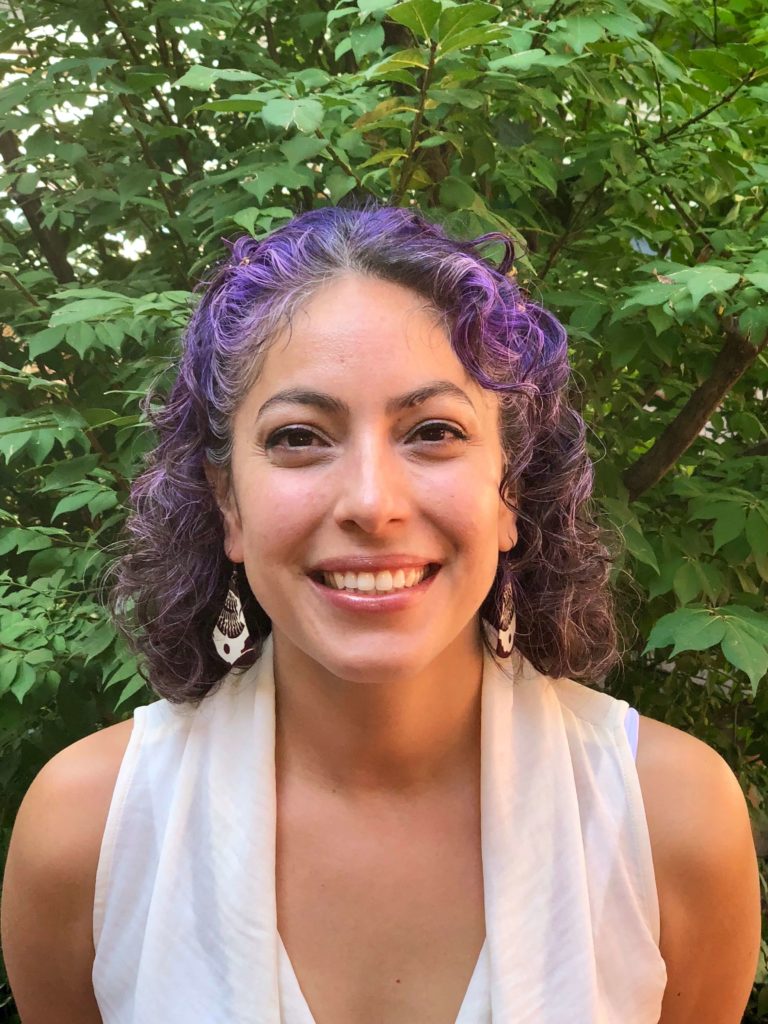 A Latina woman in a white sleeveless blouse and big dangle earrings poses in front of foliage. She has shoulder-length curly purple hair, eyeliner, and a smile that reveals her top teeth.