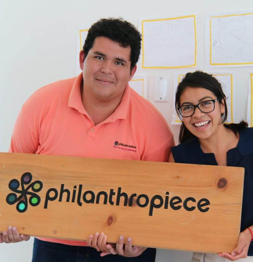 Two people smile at the camera while holding a wooden board engraved with the philanthropiece logo: six circles connected like a wheel in the center