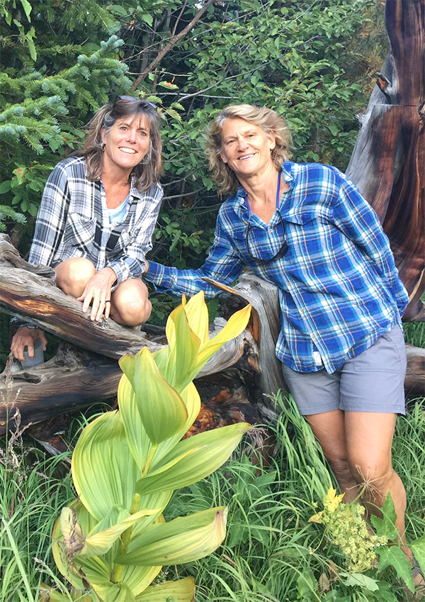Libby and Joanie, two middle-aged women, pose near a log surrounded by lush greenery