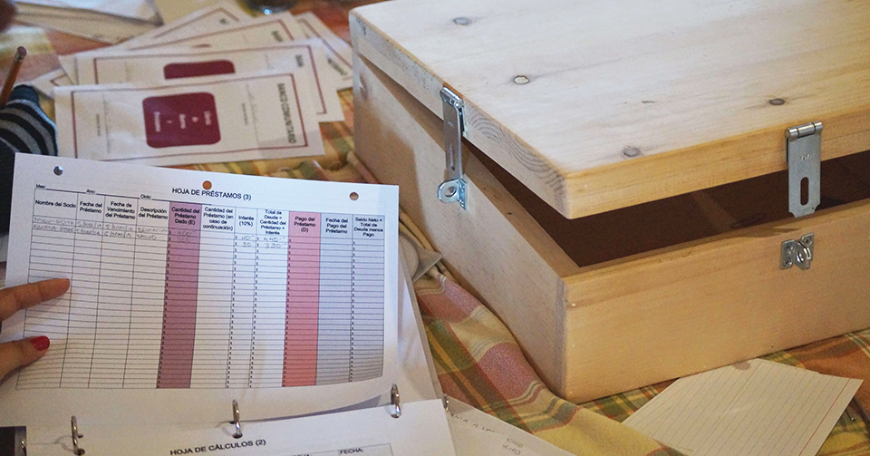 A "hoja de prestamos" or loan sheet is held up in front of a wooden box with two visible locks.