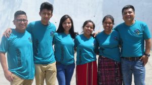 :Six people stand in a line with their arms linked. They are wearing matching blue Filantropis t-shirts.