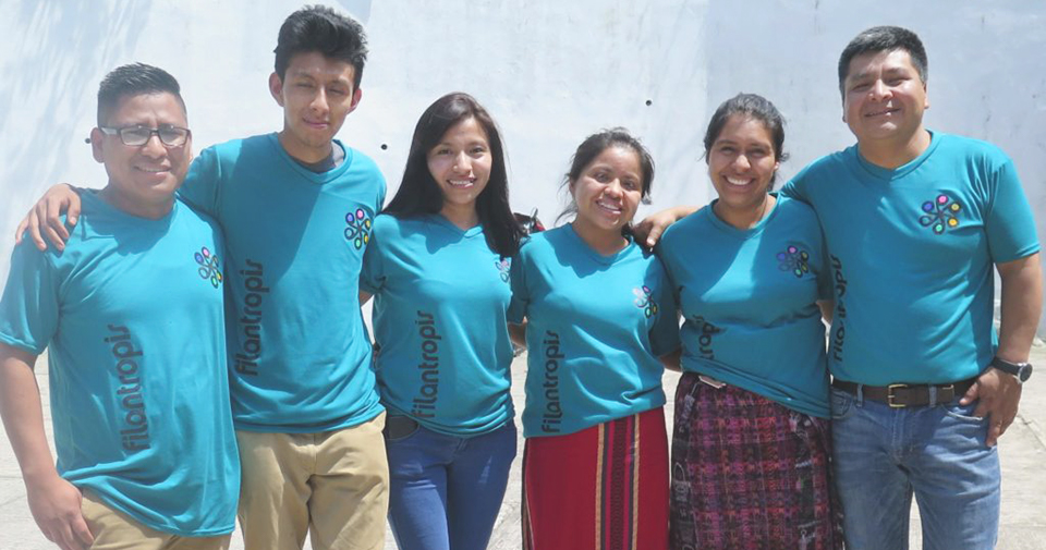 :Six people stand in a line with their arms linked. They are wearing matching blue Filantropis t-shirts.