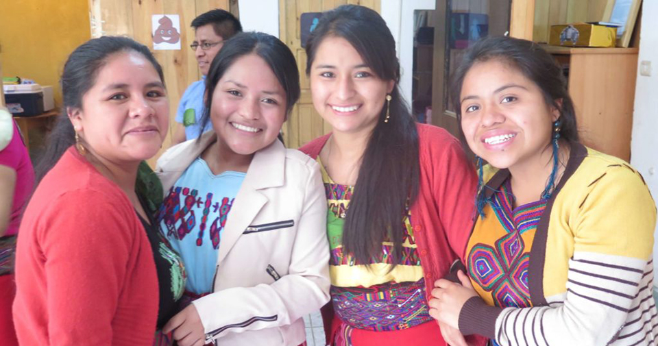 :Four women stand smiling at the camera with their arms linked. They are wearing a mix of traditional embroidered clothing and "modern" fashion like cardigans and blazers