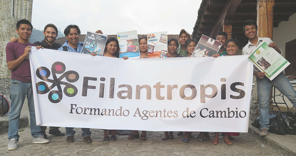 A dozen of people stand on a cobblestone road holding a banner that says "Filantropis. Formando Agentes de Cambio."