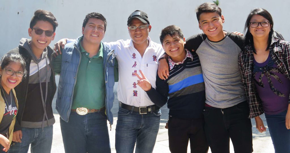 :Seven people stand in a line with their arms linked around each other. The one in the middle is holding up a peace sign, and everyone is smiling