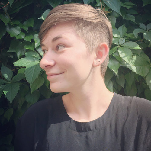 Raye poses candidly in front of green leaves, looking to their right and smiling. They have a short blonde undercut, a nose ring, earrings, and a black silk shirt.