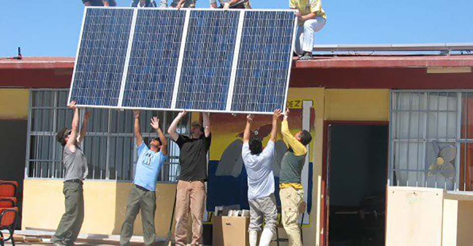 Five people are shown hoisting a solar panel up to five other people standing on the roof of a small building.