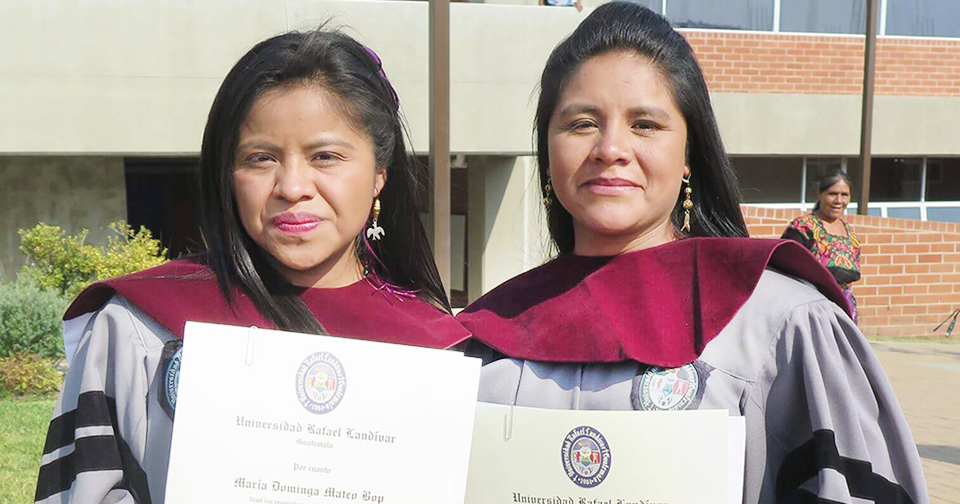 :Two women stand outside in lavender graduation gowns holding their diplomas