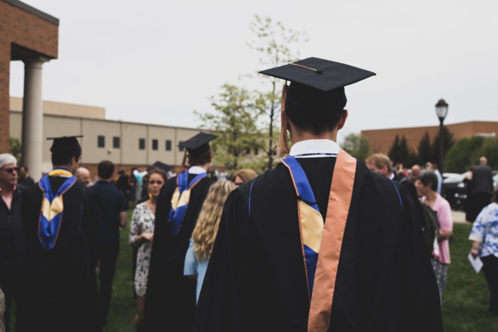 a person in a graduation cap and gown is pictured from behind. They are looking out onto a university courtyard full of milling people.