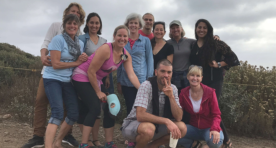 About a dozen people from the CO office cluster together, some crouched in the front row. They are smiling in front of dark clouds and plants. The photo is taken angled upwards.