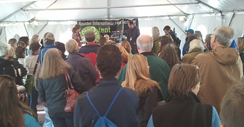 Dozens of people stand huddled under a white event tent, looking at a stage