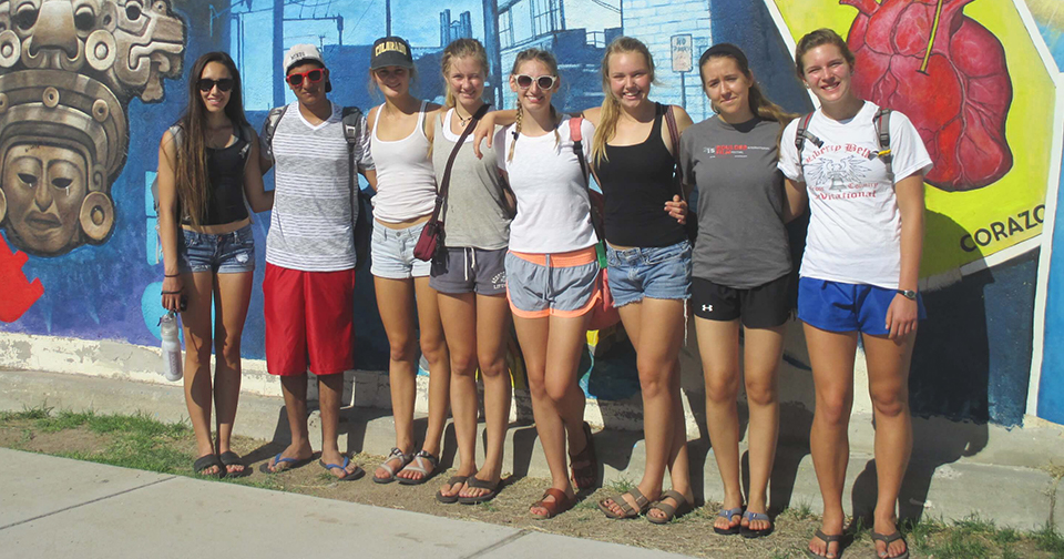 Eight teenagers stand in front of a monochrome blue mural and smile, squinting into the sun.