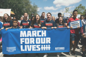 A group of teenagers hold a blue sign with white text that says "March For Our Lives." They are wearing matching navy shirts and are all mid-chant.