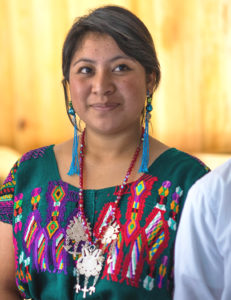 a candid of karen shows her in front of a wood-paneled wall smiling slightly into the distance. She is wearing blue tassel earrings, a beaded necklace with a large silver pendant, and a colorfully-embroidered blouse.