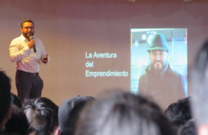 A photo taken over the heads of an audience reveals a man in a white button-up speaking into a microphone. He is accompanied by a projected PowerPoint that says "the learning journey"
