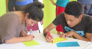 Two youth hunch over a white plastic table, intently writing on pieces of construction paper with some markers