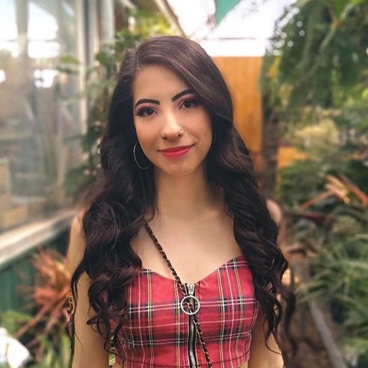 Jeny is pictured from the elbows up. She is standing in a greenhouse and wearing a plaid sleeveless shirt. Her dark hair falls to her ribs, and she is wearing bright lipstick and hoop earrings.