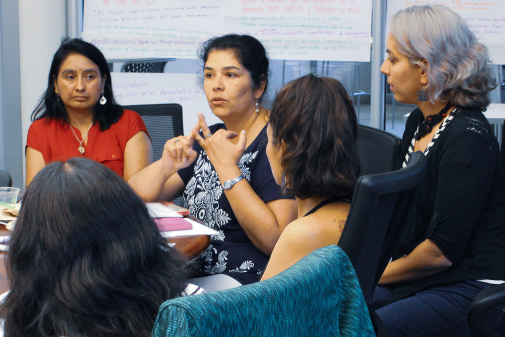 5 Latina women are pictured sitting around a table. One of them is talking, and the rest are listening intently.