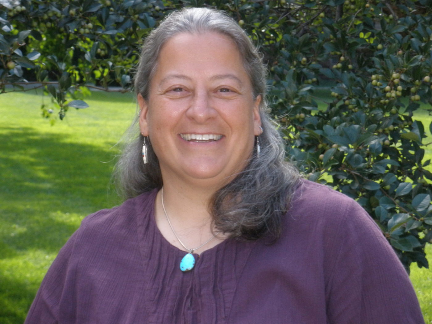 A picture of Doreen from the chest up. She is a middle-aged woman with collarbone-length gray hair and a full-toothed smile. She wears a purple blouse, dangle earrings, and a turquoise necklace.